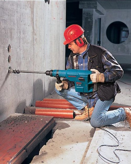 Worker in hard hat using Bosch GBH 11 DE rotary hammer to drill through concrete wall on demolition site