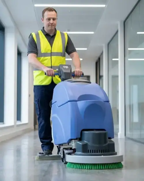 Walk-behind scrubber dryer cleaning an office corridor, green scrubbing brush at the front and squeegee drying behind
