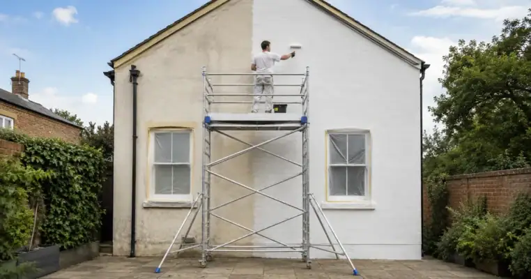 Decorator painting the gable end of a house in masonry paint from a mobile scaffold tower with the windows masked off below